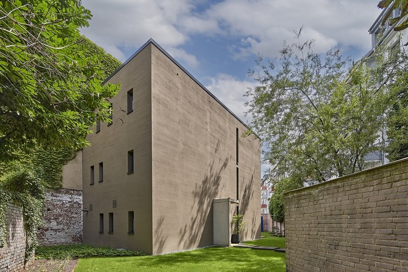 Na trudnej, pełnej zakamarków działce w samym centrum miasta udało się wzorowo rozwiązać problem ograniczonej przestrzeni i deficytu mieszkań. Od strony zachodniej, o monolitycznym, proporcjonalnym charakterze, uwagę przyciąga prostota – jedynie drzwi wejściowe i wąskie szczeliny świetlne przerywają gładką powierzchnię.